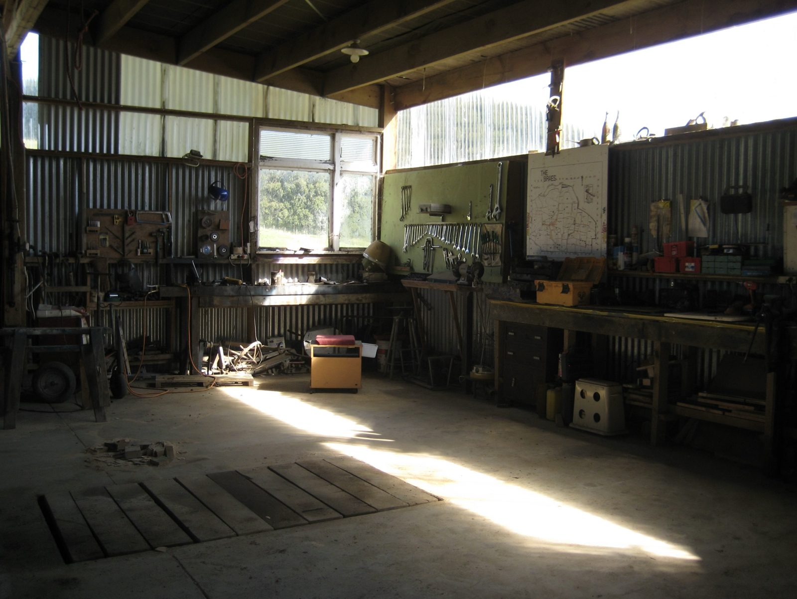 Inside a corrugated-iron farm workshop: spanners on a pegboard, workbenches, a hand-drawn farm map on the wall, and sunlight across a concrete floor with a timber-covered service pit